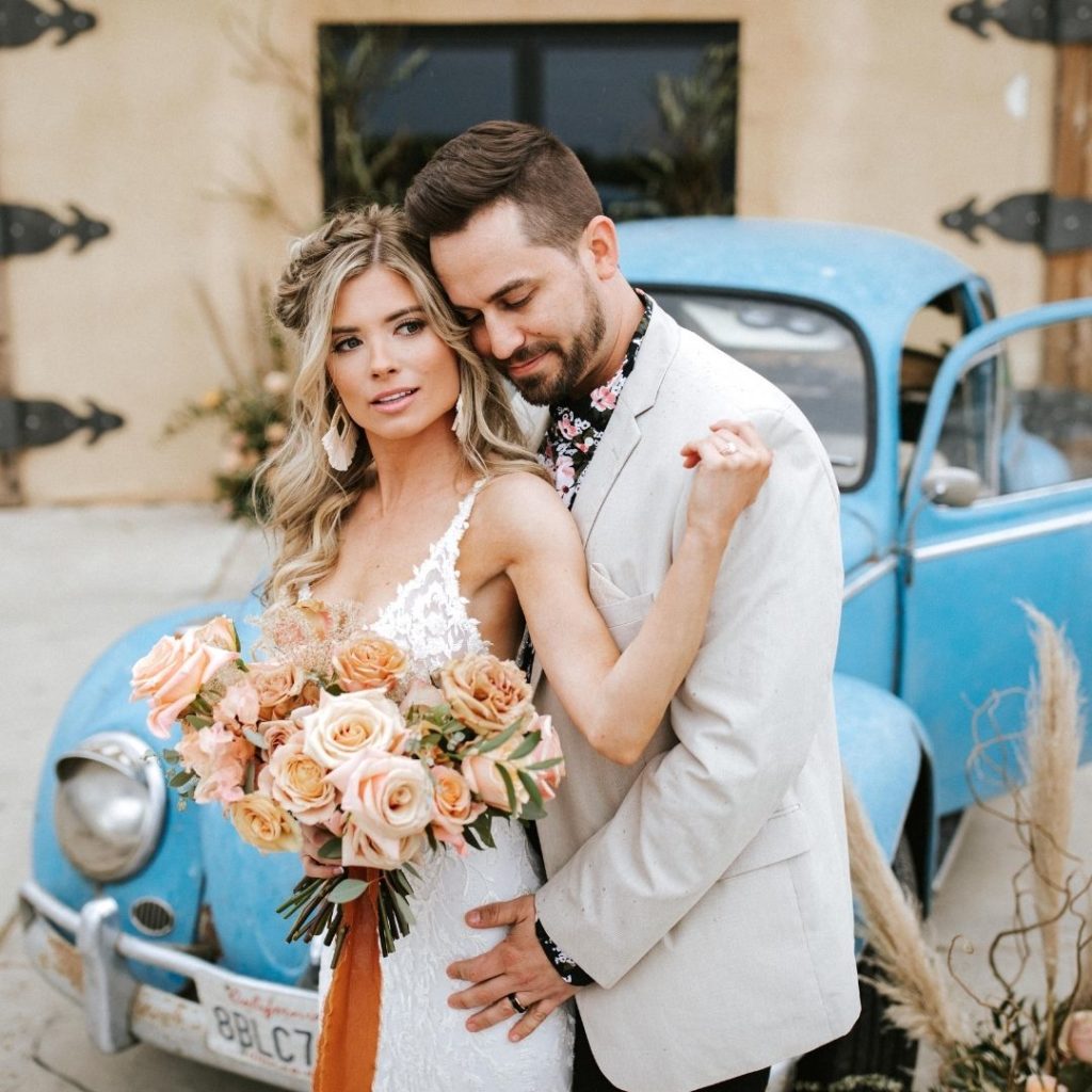 Shows a bride and groom with boho chic style posing in front of a baby blue Volkswagen Beetle.