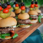 Display showing gourmet burger sliders with bacon and fresh greens topped with a toothpick skewered with a pickle, baby tomato, and black olive